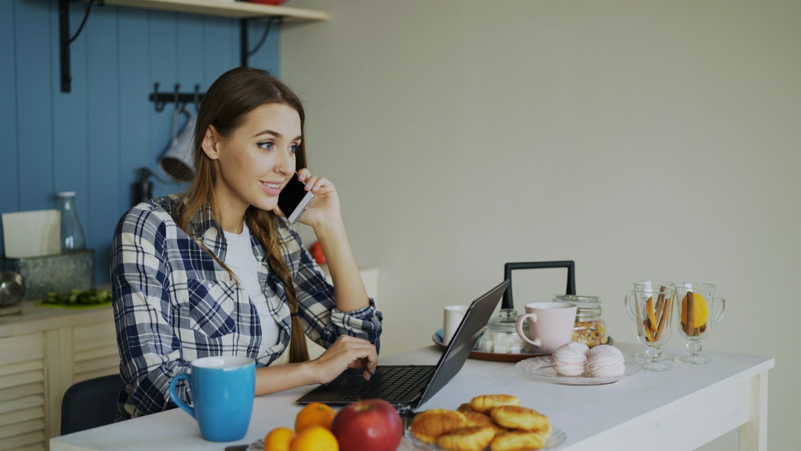 A woman at a kitchen table on the phone while working on a laptop, the everyday setting in which parents and staff receive a school's update about a cyber incident.